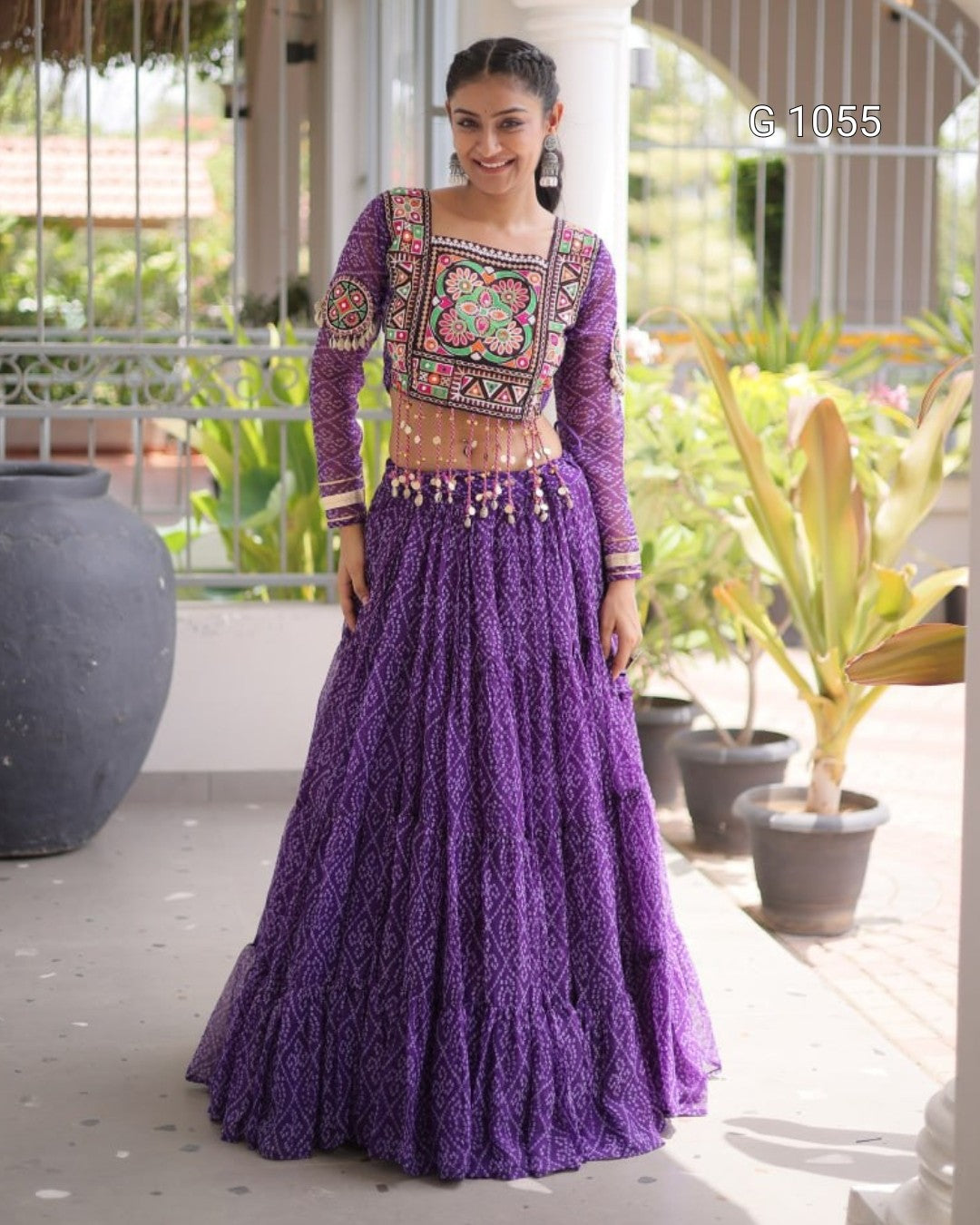 A woman models a traditional Indian ethnic wear set consisting of a purple embroidered lehenga skirt and a matching long-sleeve blouse with intricate geometric and floral embroidery. The set is photographed outdoors on a covered patio with potted plants in the background.