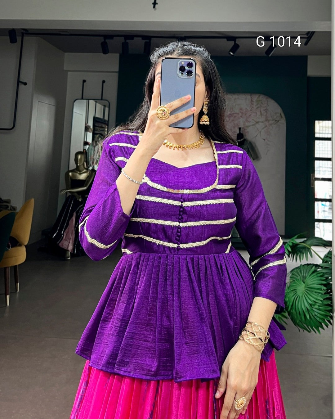 A woman takes a mirror selfie wearing a purple traditional-style top with white horizontal stripes and a bright pink skirt. She is holding a blue smartphone in front of her face, with jewelry on her hands and arms.