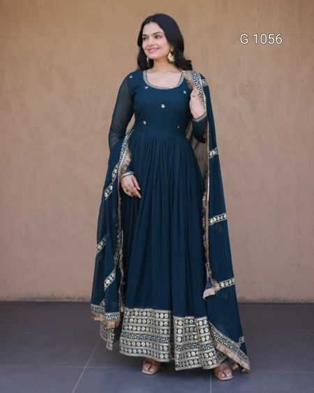 A woman modeling a dark teal blue anarkali gown with gold embroidery and a matching dupatta, standing against a textured beige wall.