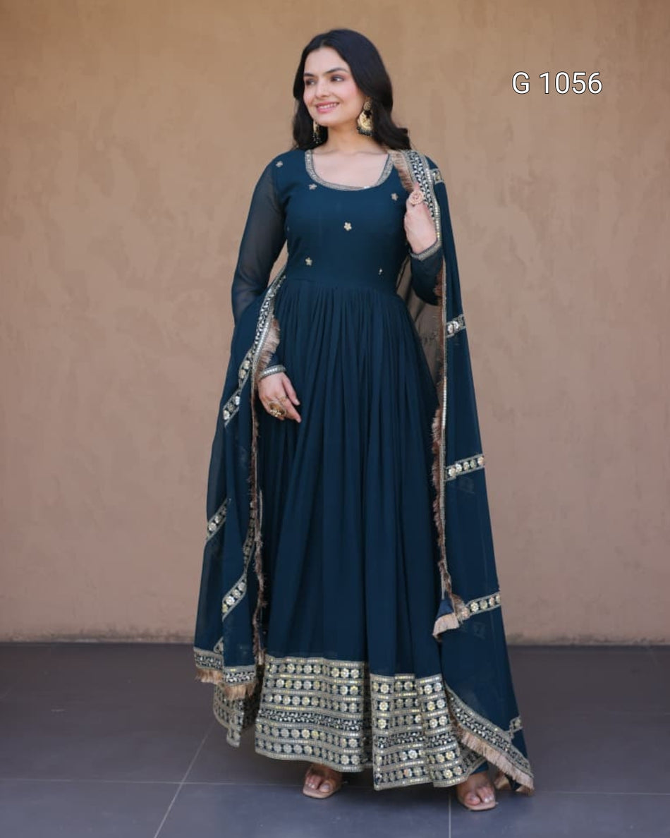 A woman modeling a dark teal blue anarkali gown with gold embroidery and a matching dupatta, standing against a textured beige wall.