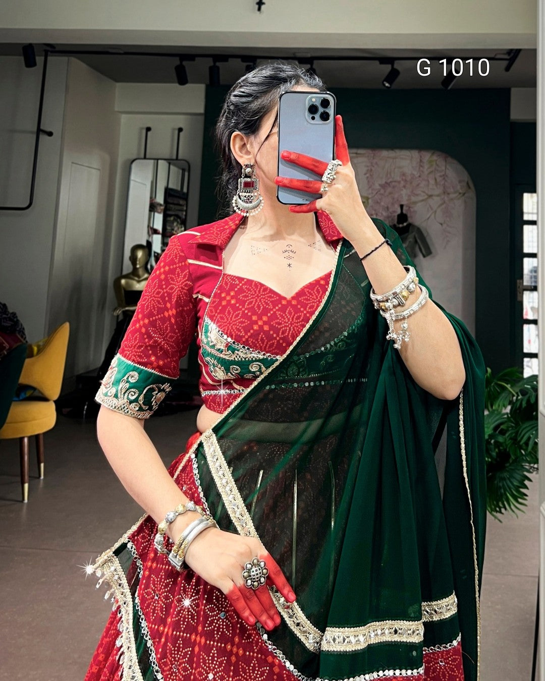 A woman takes a mirror selfie wearing a traditional Indian outfit consisting of a red and green patterned blouse, a matching red skirt, and a dark green dupatta with silver borders. She is wearing heavy silver jewelry and holding a smartphone to take the photo.