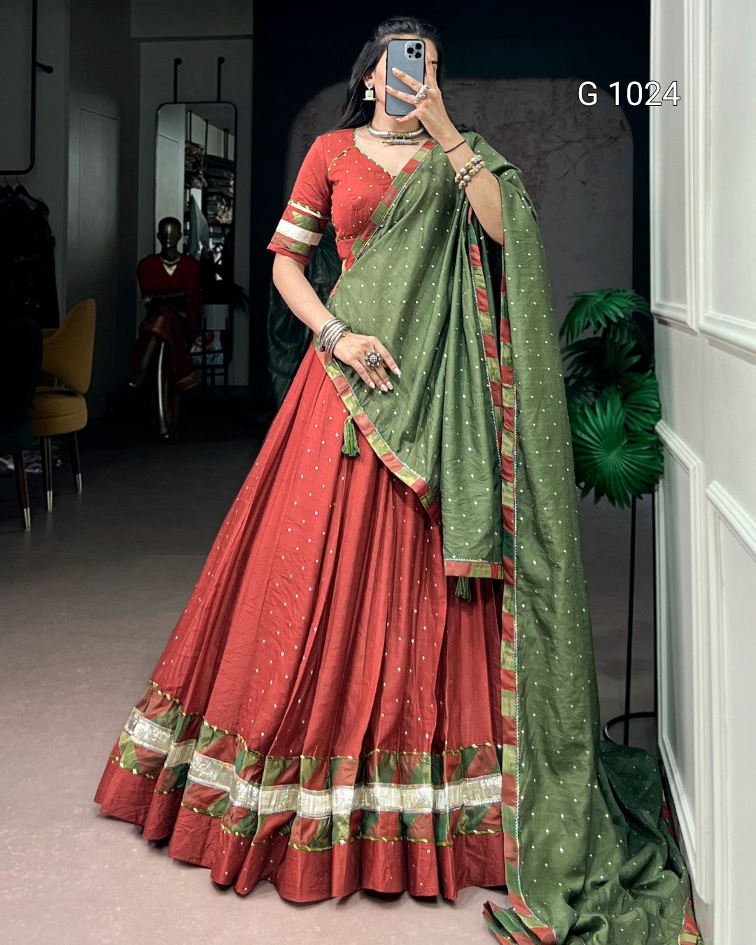 A woman wearing a traditional Indian outfit consisting of a rust-red flared skirt (lehenga) with green and gold border details, a matching rust-red short-sleeve top, and a green dupatta with gold motifs, taking a mirror selfie in an indoor setting.