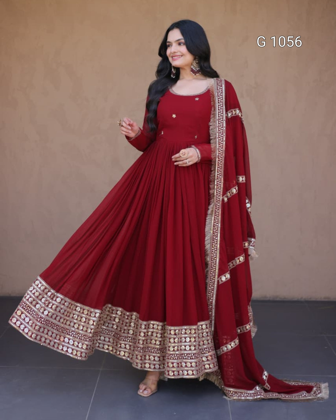 A woman models a deep red Indian ethnic gown (Anarkali) with a matching dupatta, featuring intricate gold and silver mirror work borders. The background is a plain beige wall with a gray tiled floor, and the text 'G 1056' is visible in the top right corner.