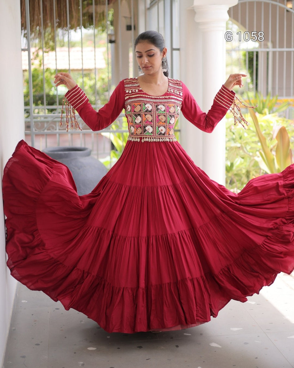 A woman wearing a long-sleeved red Anarkali gown with intricate embroidery on the bodice and sleeves, twirling to show off the full tiered skirt in an outdoor porch setting.