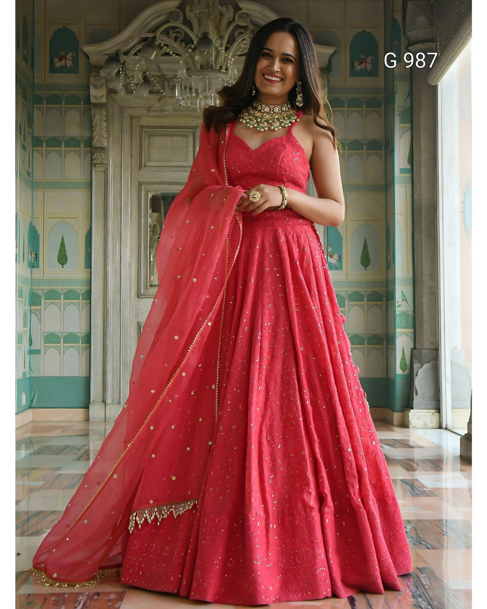 A woman models a vibrant red lehenga choli gown with gold embroidery and a matching dupatta in an ornate indoor palace setting. The image includes a style code 'G 987' in the top right corner.