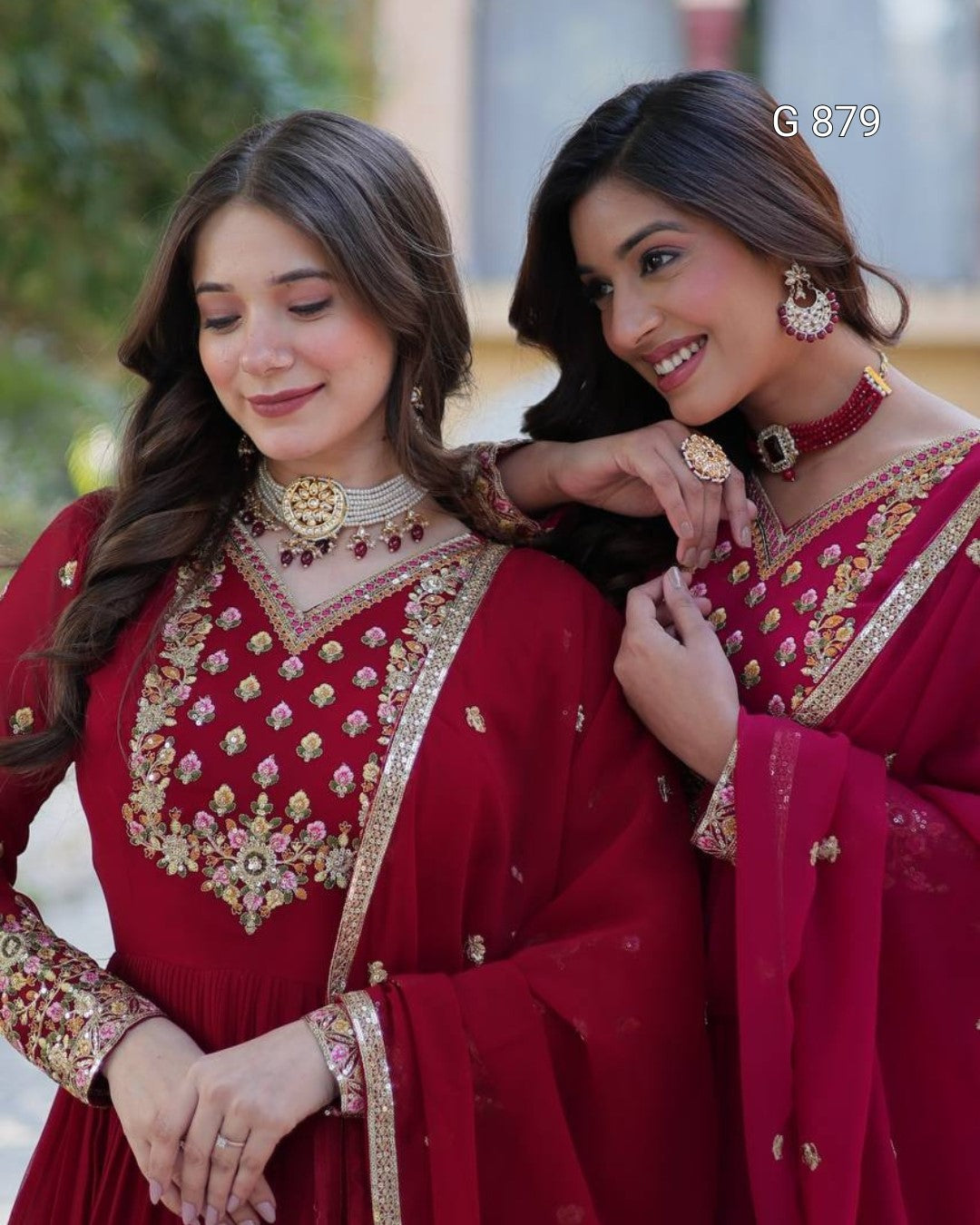 Two women in traditional red embroidered dresses with gold detailing, standing close together outdoors.