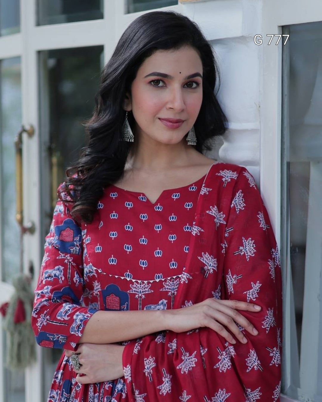 Woman wearing a red dress with blue patterns standing against a white wall.