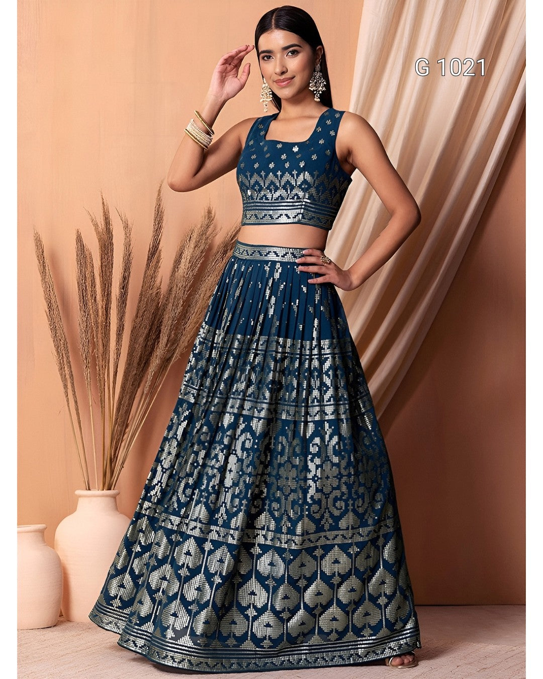 A woman models a dark blue and silver printed traditional Indian outfit consisting of a sleeveless crop top and a long flared skirt, set against a warm beige backdrop with decorative dried grass in a vase.