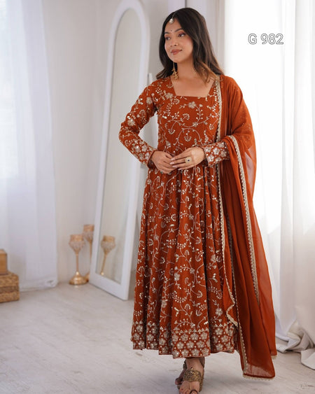 A woman models a rust-orange Anarkali gown with intricate gold floral embroidery and a matching dupatta in a bright, white-walled room.
