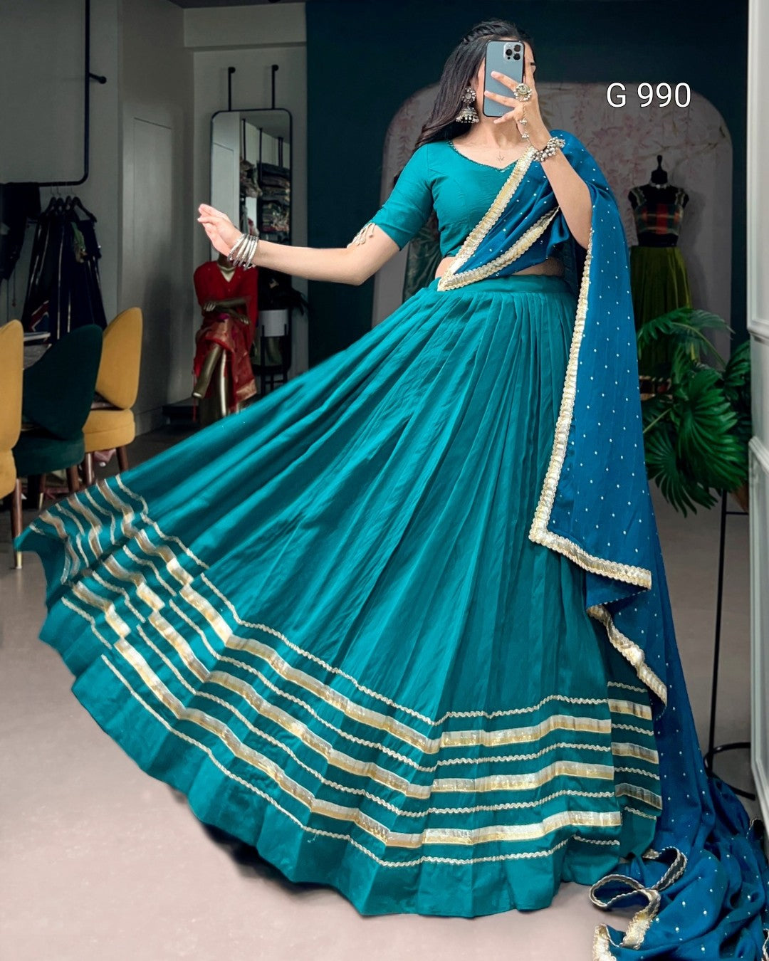 A woman modeling a teal blue traditional Indian lehenga choli dress with gold zari stripes, holding a phone to take a mirror selfie in a boutique setting.