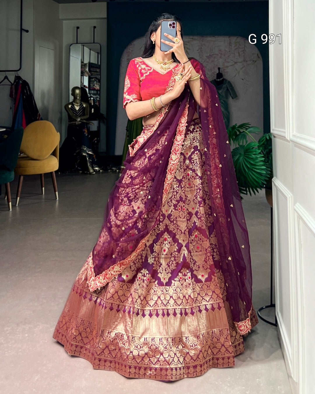A woman takes a mirror selfie wearing an ornate Indian ethnic lehenga choli. The outfit features a deep purple embroidered skirt (lehenga) with intricate gold and pink metallic patterns, paired with a bright pink blouse.