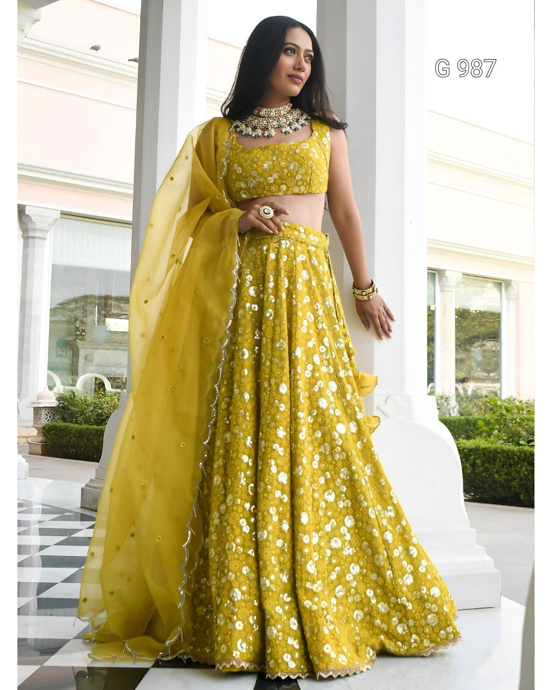 A woman models a bright yellow embroidered Indian ethnic outfit consisting of a sleeveless crop top, a flared skirt, and a matching sheer dupatta, set against a white architectural background.