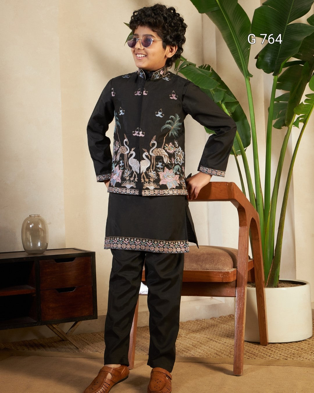 Young boy in a traditional outfit standing in a room with a plant and wooden chair.
