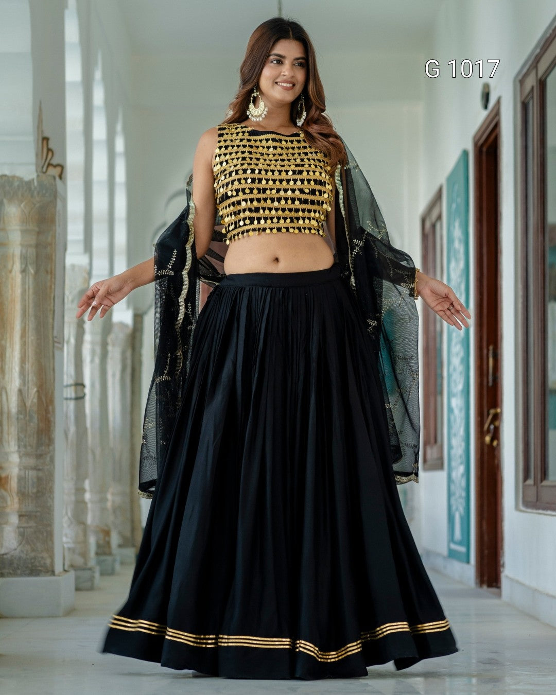 A woman modeling a black and gold traditional Indian outfit with a sleeveless gold-embroidered crop top, a full black skirt with gold border stripes, and a sheer black dupatta, standing in a hallway.