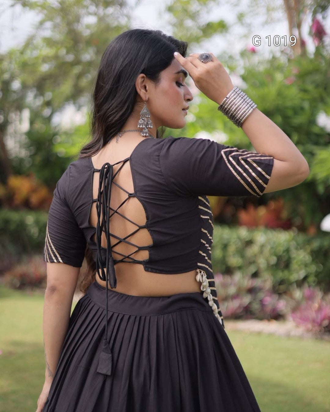 A woman wearing a black short-sleeve blouse with an open lace-up back and striped sleeve details, paired with a black pleated skirt, photographed outdoors in a garden setting.
