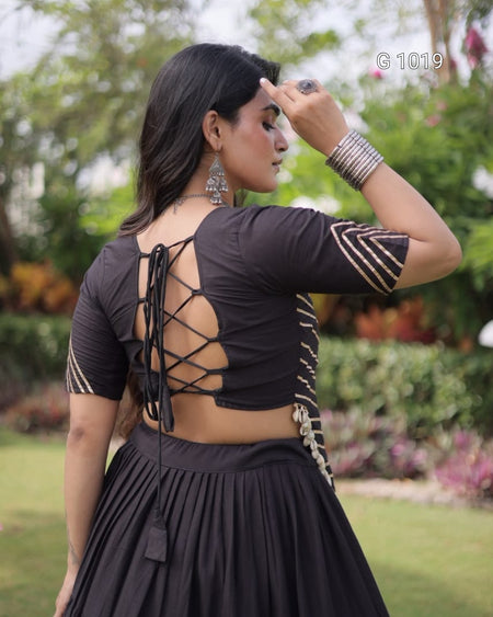 A woman wearing a black short-sleeve blouse with an open lace-up back and striped sleeve details, paired with a black pleated skirt, photographed outdoors in a garden setting.
