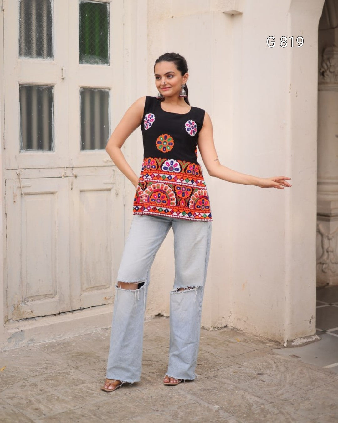 Woman wearing a black sleeveless top with colorful patterns and light blue jeans standing in front of a white wall.