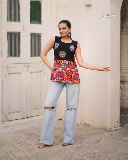 Woman wearing a black sleeveless top with colorful patterns and light blue jeans standing in front of a white wall.