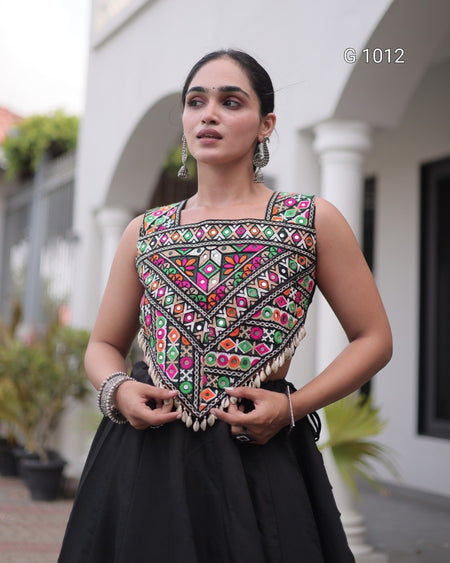 A woman wearing a sleeveless black skirted dress with a highly detailed, colorful geometric embroidered bodice and fringe, standing outdoors in front of a white building with arches.