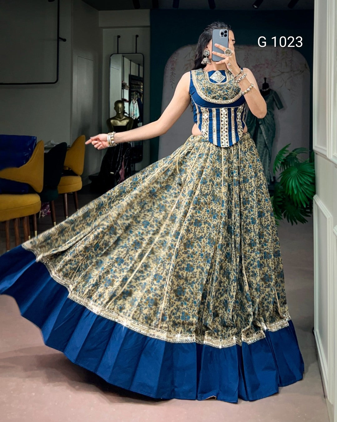 A woman takes a mirror selfie in a boutique wearing a traditional Indian anarkali gown. The dress features a blue and gold floral printed skirt with a solid royal blue pleated border, and a blue sleeveless bodice with gold embroidery.