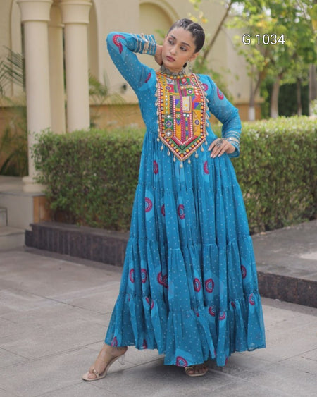 A woman modeling a long-sleeved blue traditional dress with colorful embroidered details on the chest and scattered circular motifs on the skirt, posed outdoors in front of a building with columns and hedges. The text 'G-1034' is visible in the top right.