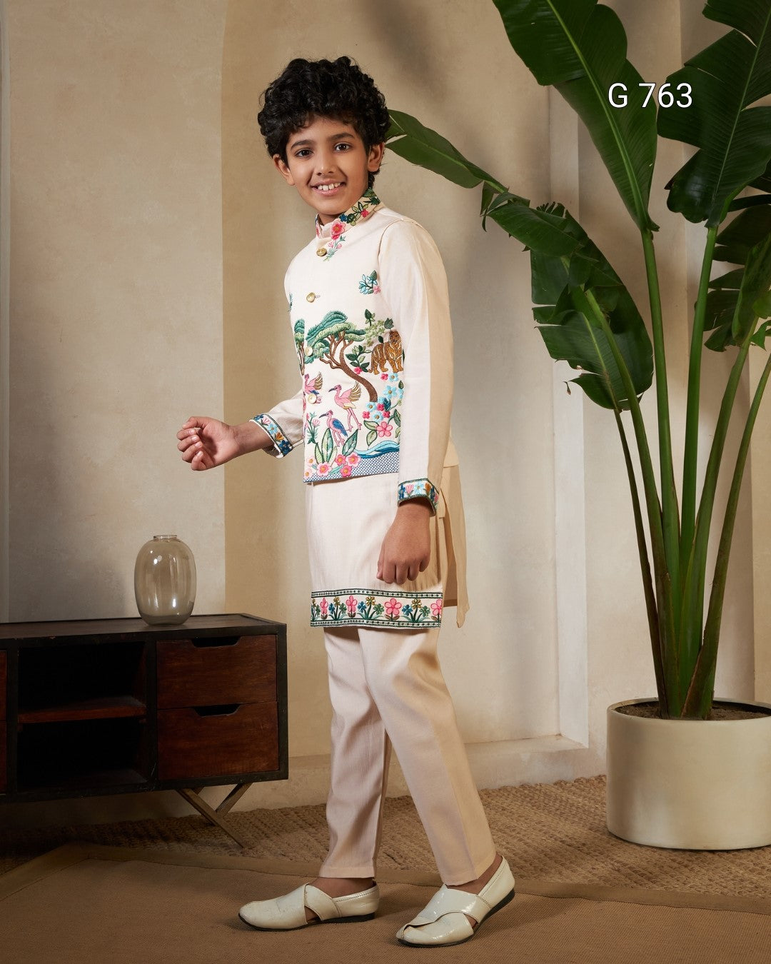 Young boy in traditional outfit standing in a room with a plant and wooden cabinet.