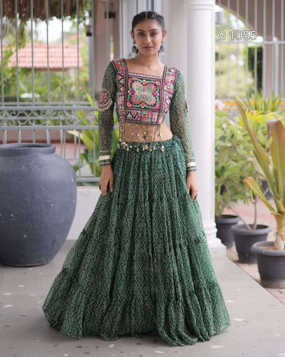 A woman modeling a dark green embroidered traditional Indian outfit with a heavily patterned embroidered bodice and a matching flared skirt, standing in a porch setting with potted plants.