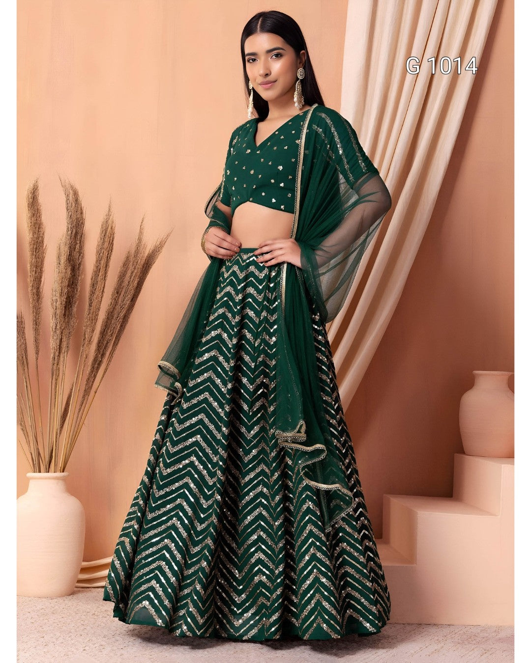 A woman models a dark green traditional Indian outfit consisting of a sleeveless crop blouse with silver embellishments, a matching flared skirt with a silver chevron pattern, and a sheer dupatta draped over her shoulders. The background is a warm beige studio setting with decorative dried grass in a vase and draped curtains.
