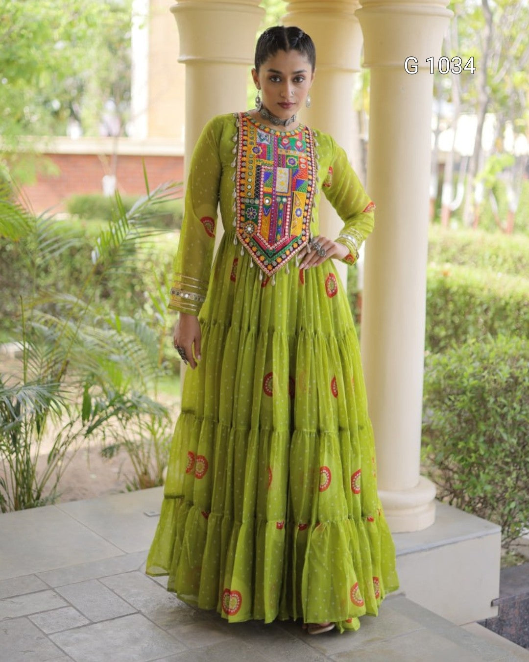 A woman models a vibrant lime green Anarkali gown with a heavily embroidered multicolor yoke and tiered skirt, standing in front of cream-colored pillars and green garden foliage.