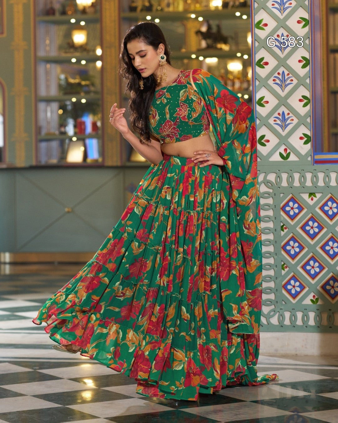 A woman models a green floral lehenga choli set with a matching dupatta in an ornate indoor setting. The outfit features a vibrant green base with red, pink, and yellow floral patterns, paired with a matching embellished blouse.
