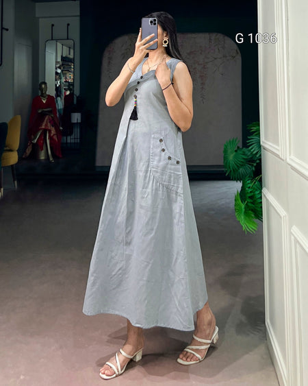 A woman takes a mirror selfie wearing a sleeveless light blue linen midi dress with button details down the front and on the pocket. The setting is an indoor boutique with a gray floor, white paneled wall, and a mannequin in a red dress in the background.