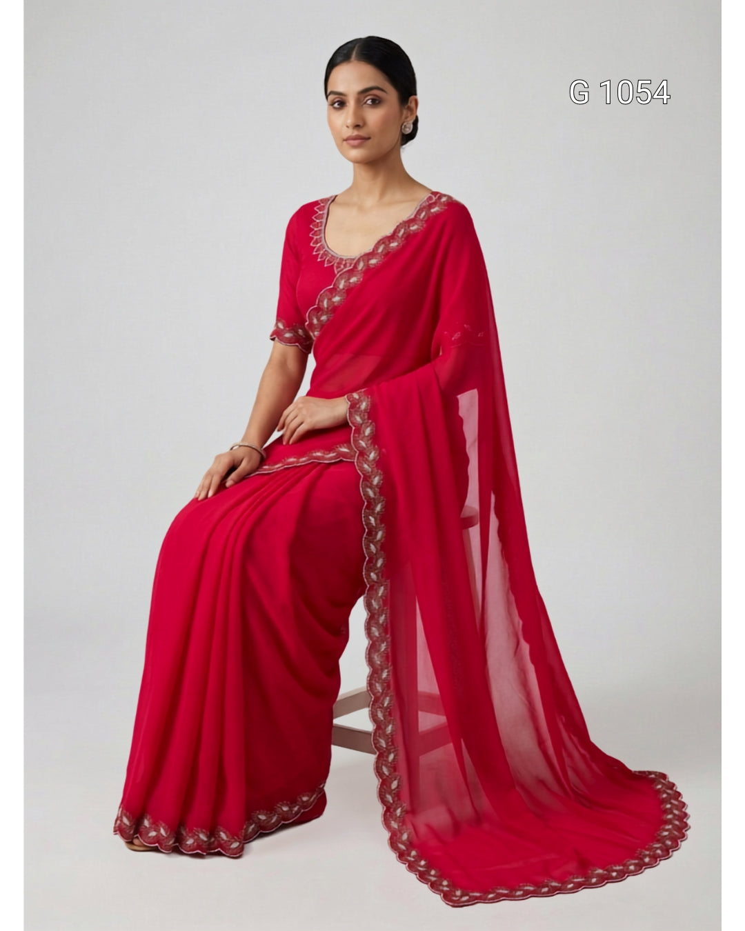 A woman seated on a stool wearing a bright red sari with metallic embroidered borders. The sari has a matching sheer dupatta draped over her lap and a short-sleeve blouse.
