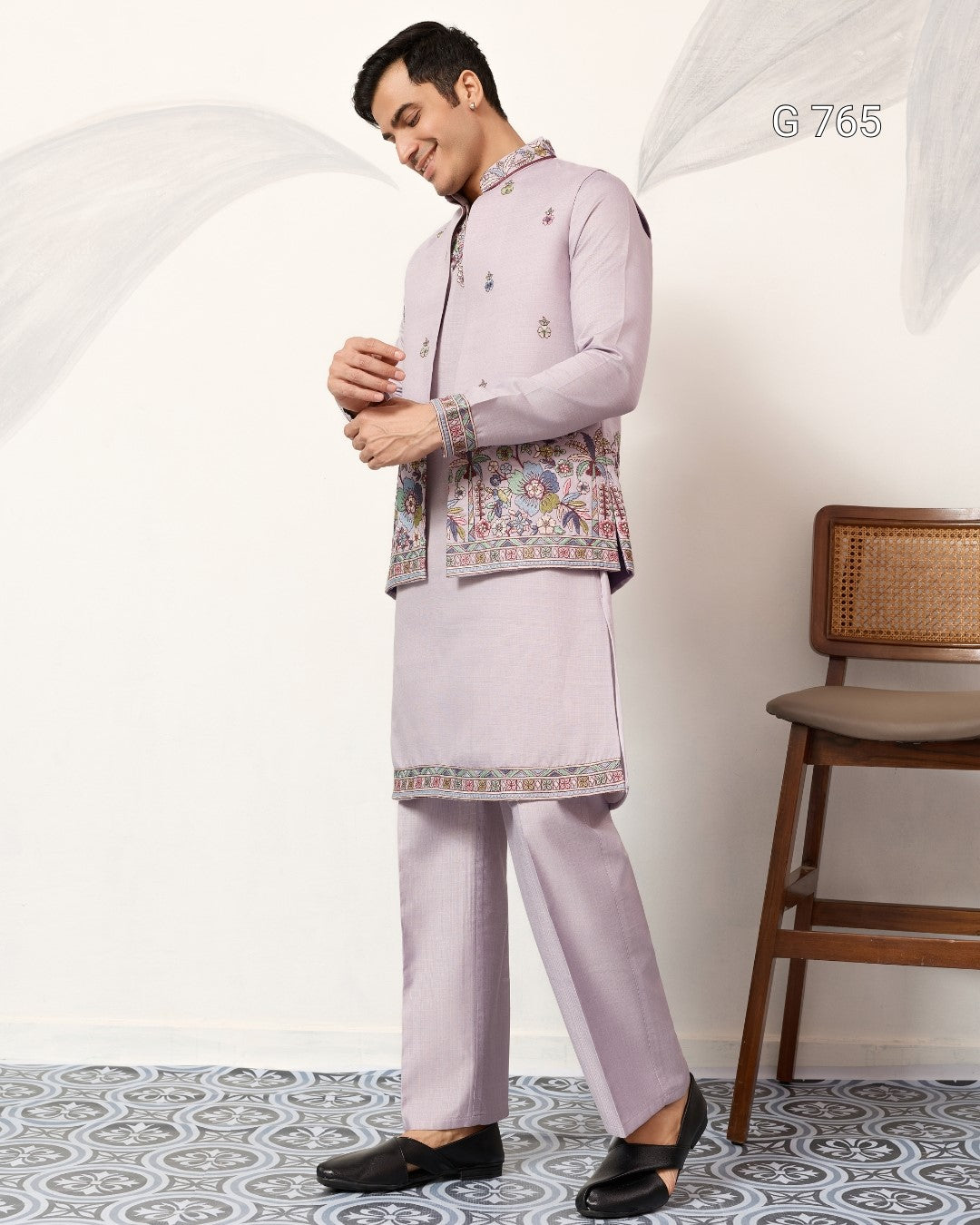 Man wearing a light purple traditional outfit with a floral jacket in a room with a chair.