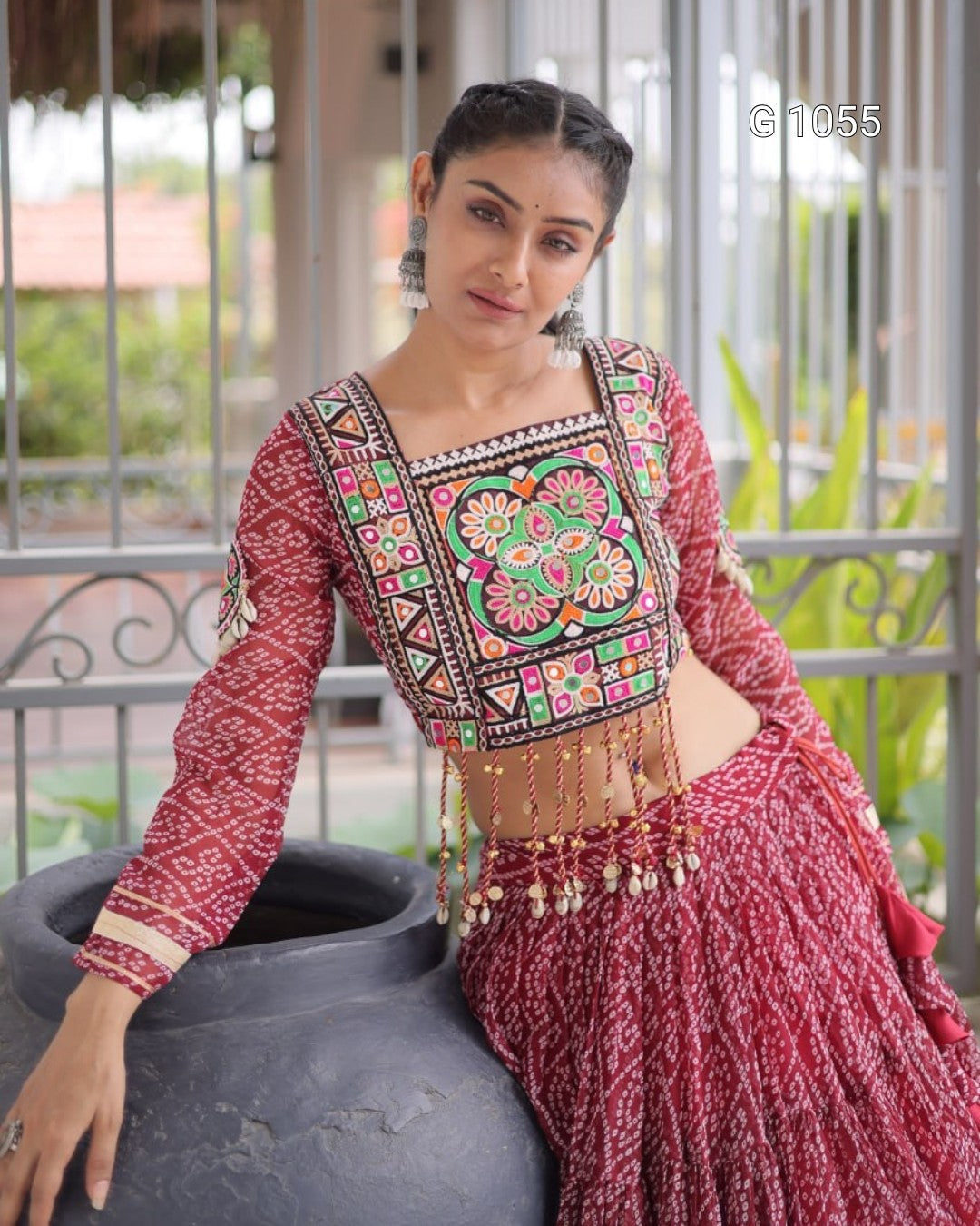 A woman models a traditional Indian ethnic wear set featuring a red patterned kurta with a heavily embroidered multicolor bodice and a matching pleated red skirt, posed outdoors next to a large dark grey pot.