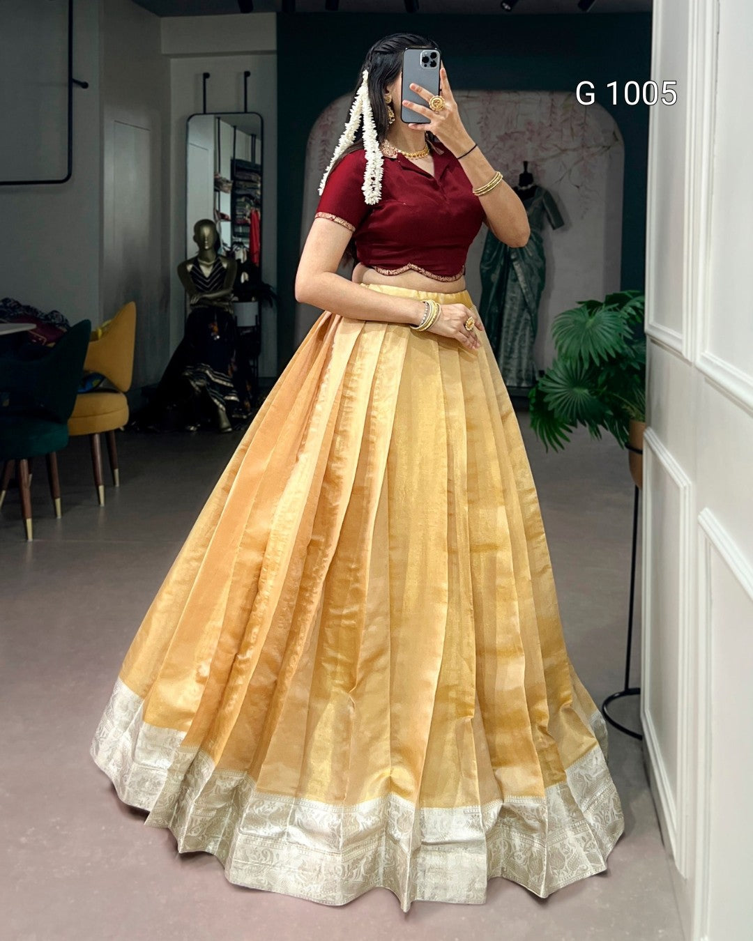 A woman takes a mirror selfie wearing a traditional Indian outfit consisting of a maroon short-sleeve blouse and a voluminous golden-yellow flared skirt with silver zari borders. The setting appears to be a boutique or dressing room interior.