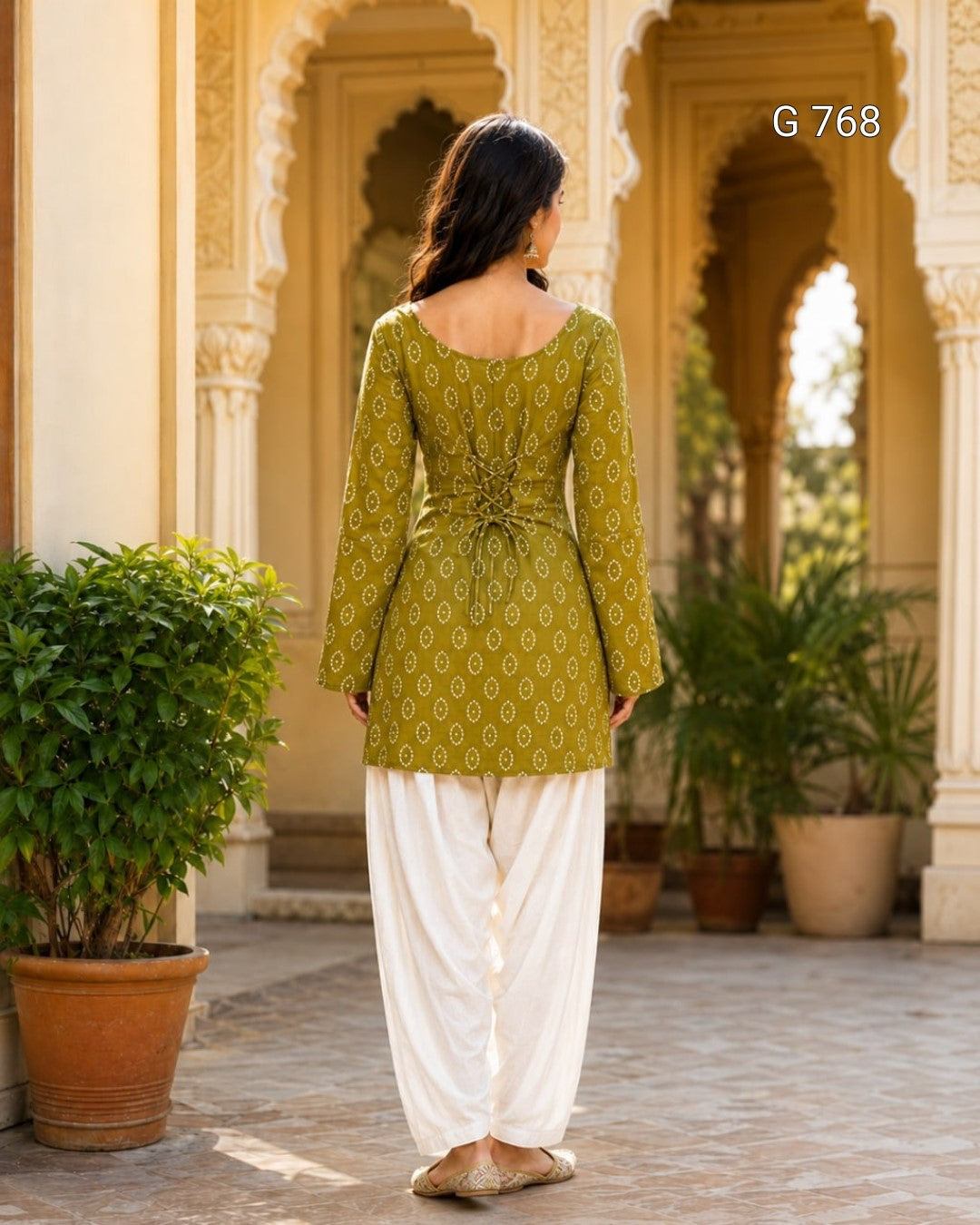Woman wearing a green patterned top and white pants in an outdoor setting with architectural elements and plants.