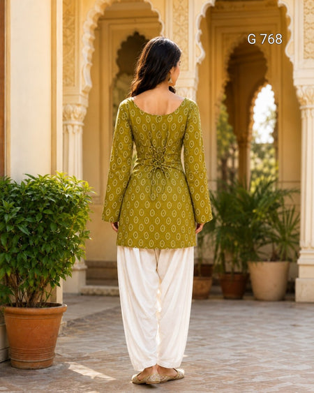 Woman wearing a green patterned top and white pants in an outdoor setting with architectural elements and plants.
