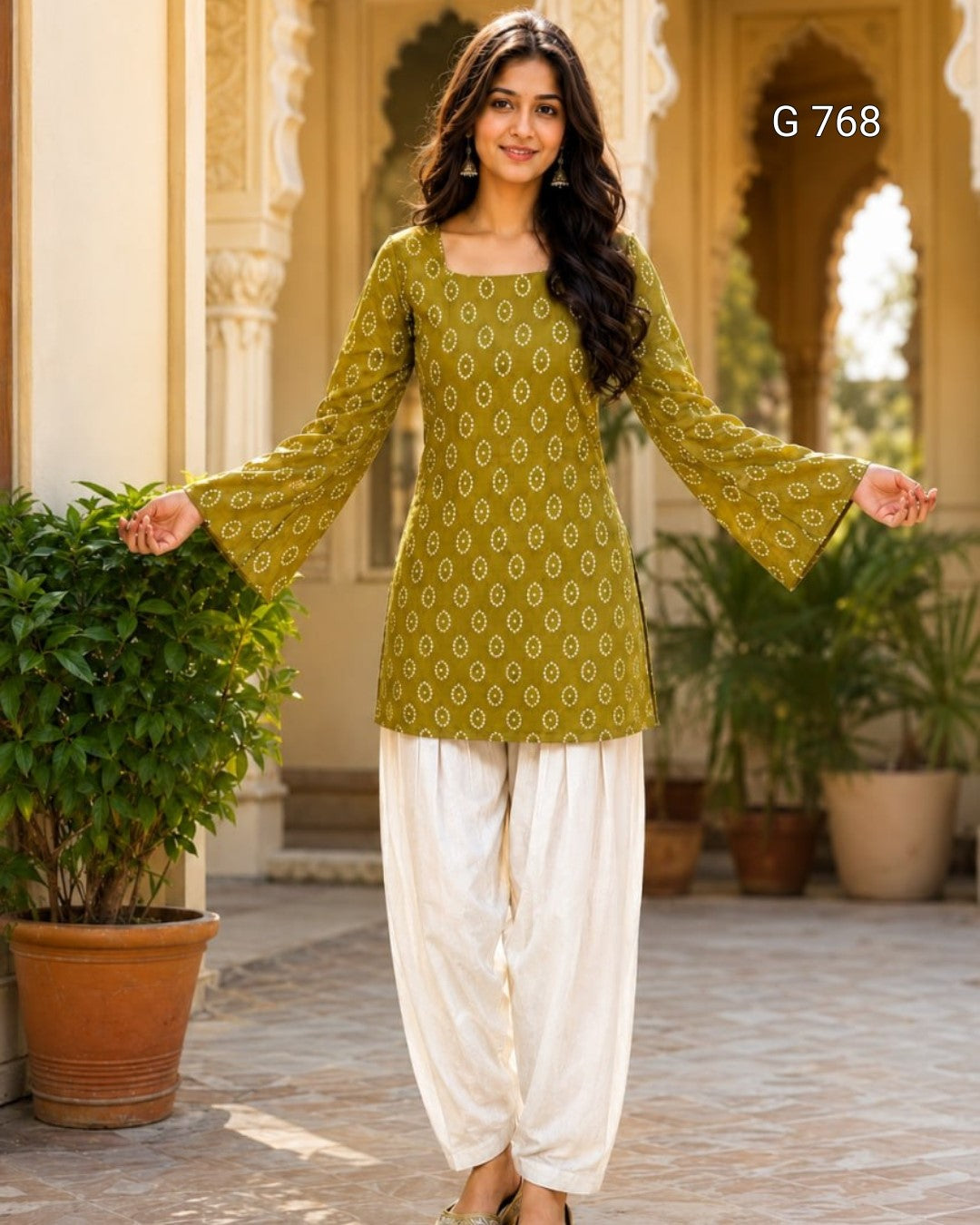 Woman wearing a green patterned top and white pants standing in an outdoor setting with plants and arches.
