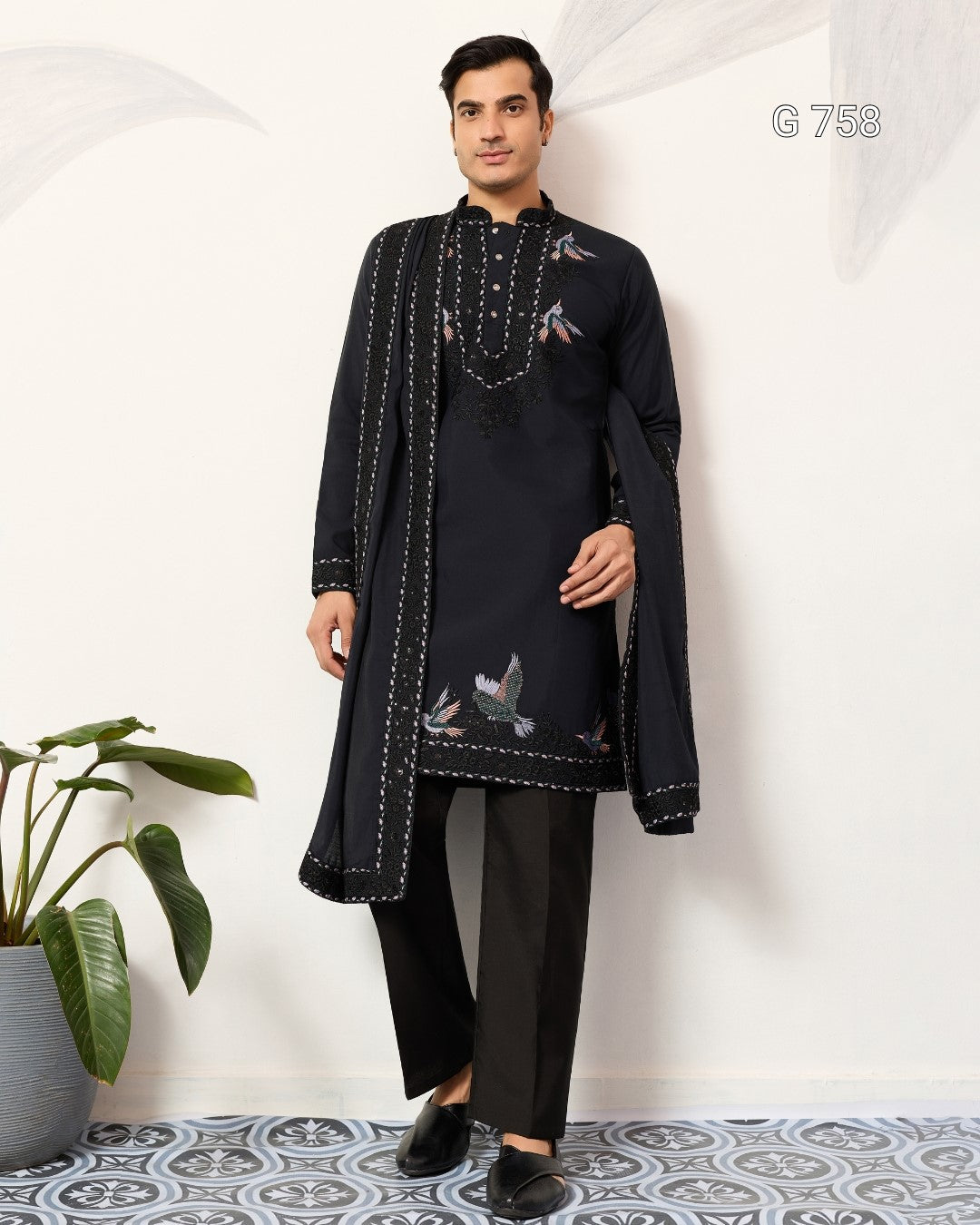Man wearing a Navy Blue embroidered traditional outfit standing against a white wall with a plant on the left.
