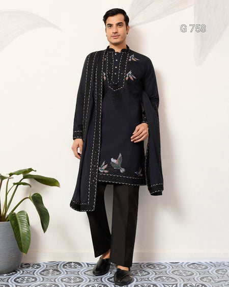 Man wearing a Navy Blue embroidered traditional outfit standing against a white wall with a plant on the left.