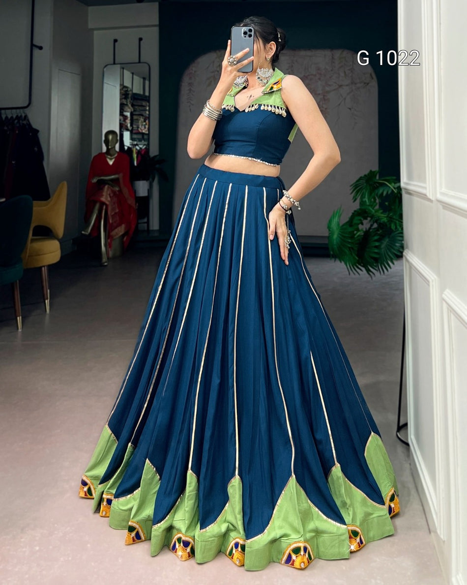 A woman takes a mirror selfie in a boutique wearing a two-piece traditional Indian outfit. The set features a dark teal blue crop top with green and gold embroidery and a matching flared skirt with vertical gold stripes and a green patterned hem.