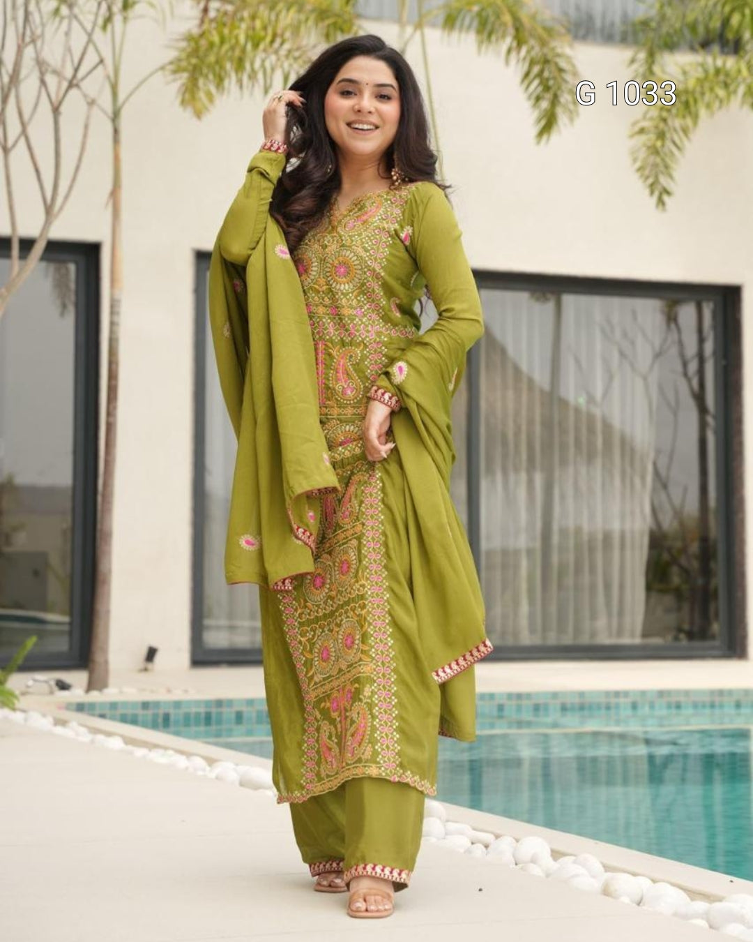 A woman models a traditional olive green 3-piece salwar kameez suit with intricate gold and pink embroidery, standing on a patio next to a swimming pool.