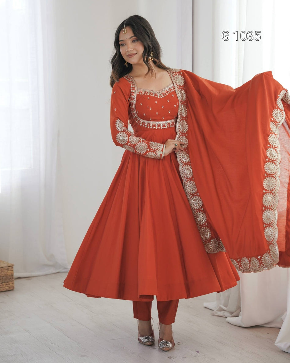 A woman modeling a traditional Indian outfit consisting of a rust-orange Anarkali kurta with gold embroidery, matching pants, and a coordinating dupatta draped over her shoulder, set against a soft white curtain background.