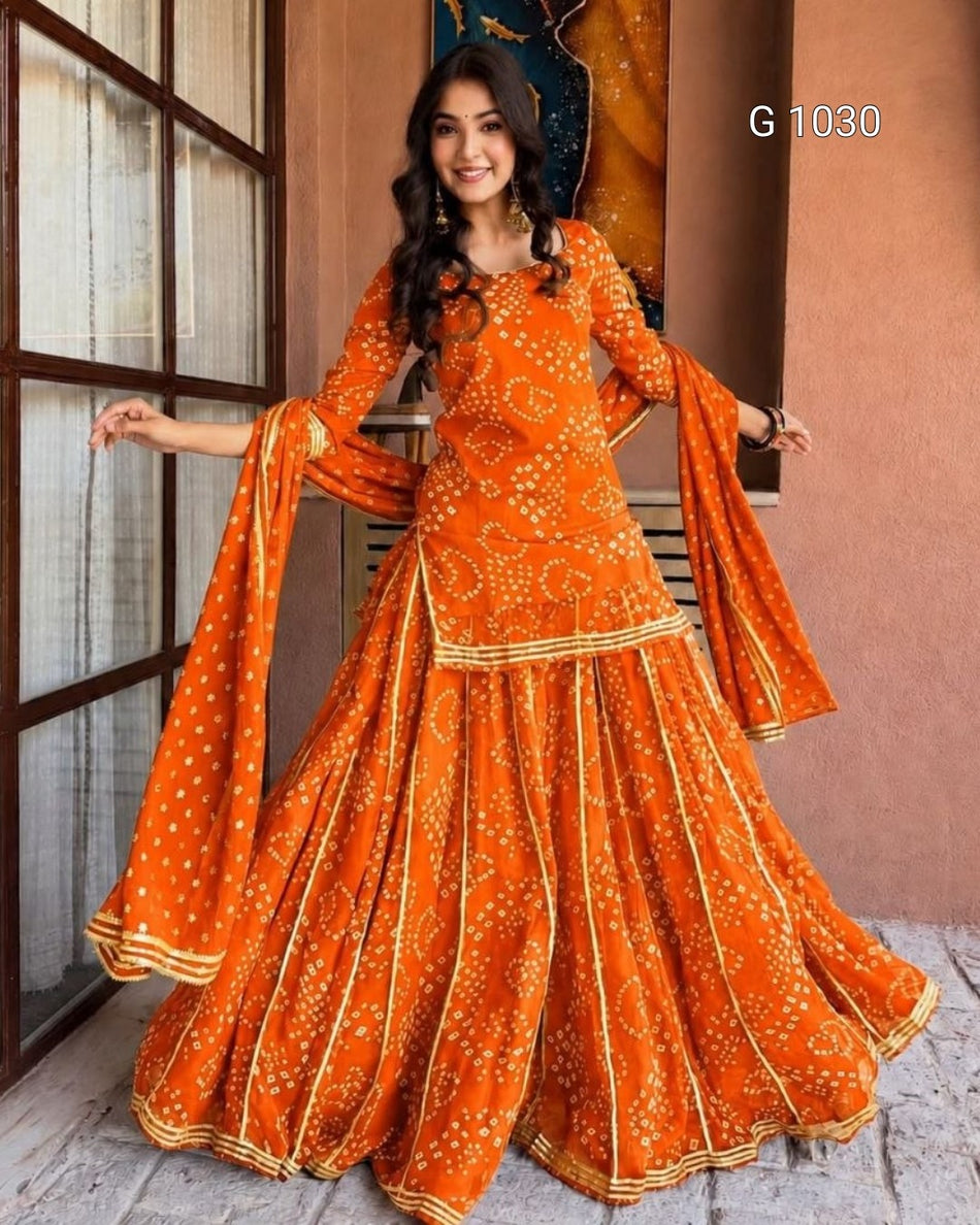 A woman modeling a bright orange traditional Indian outfit with white floral prints and gold border details, standing in front of a warm-toned wall and window frame. The text 'G 1030' appears in the top right.