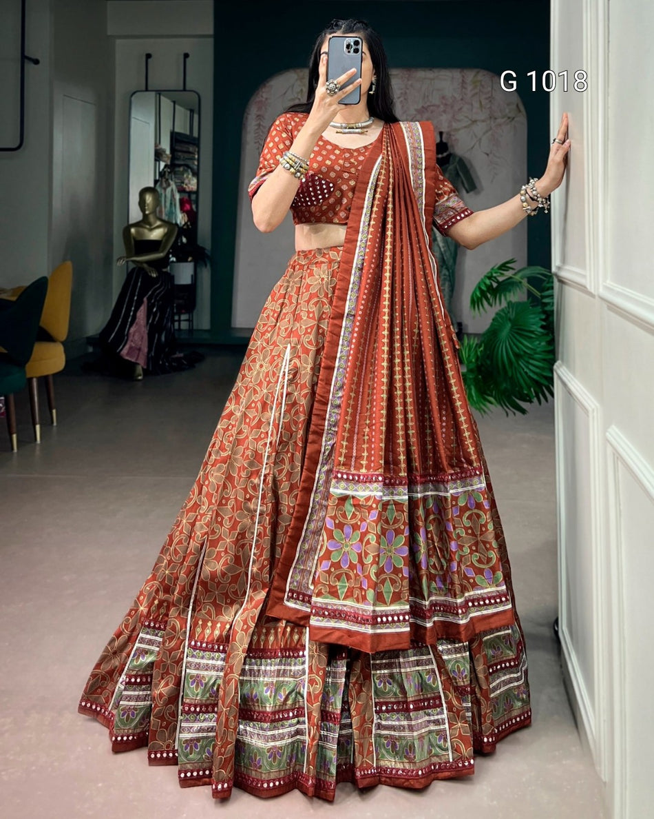 A woman takes a mirror selfie wearing a traditional Indian lehenga choli set in rust orange and brown with intricate floral patterns and a matching dupatta. The setting is an indoor boutique with a white paneled wall on the right and a mannequin visible in the background.