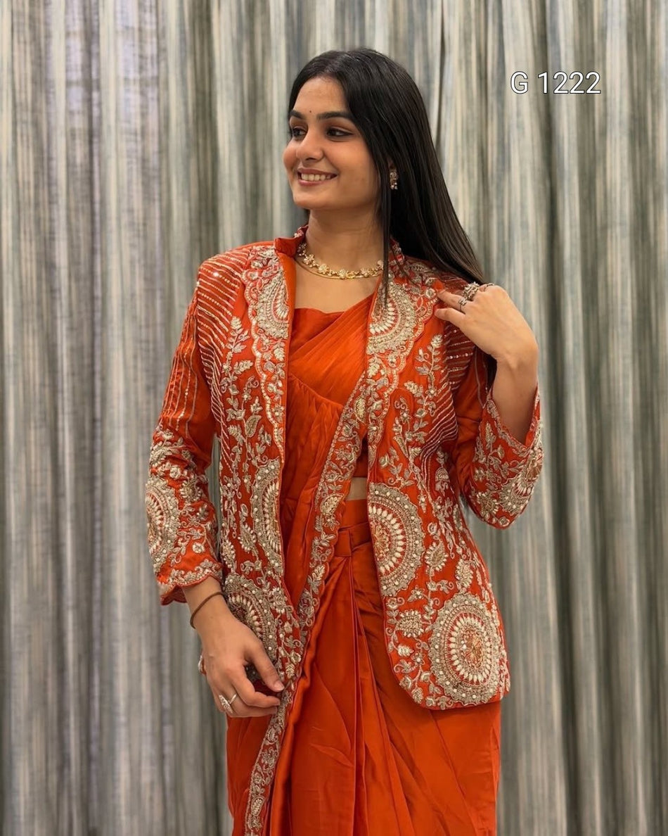 A woman wearing a bright orange saree with a matching embroidered blazer, standing in front of a gray textured curtain backdrop.