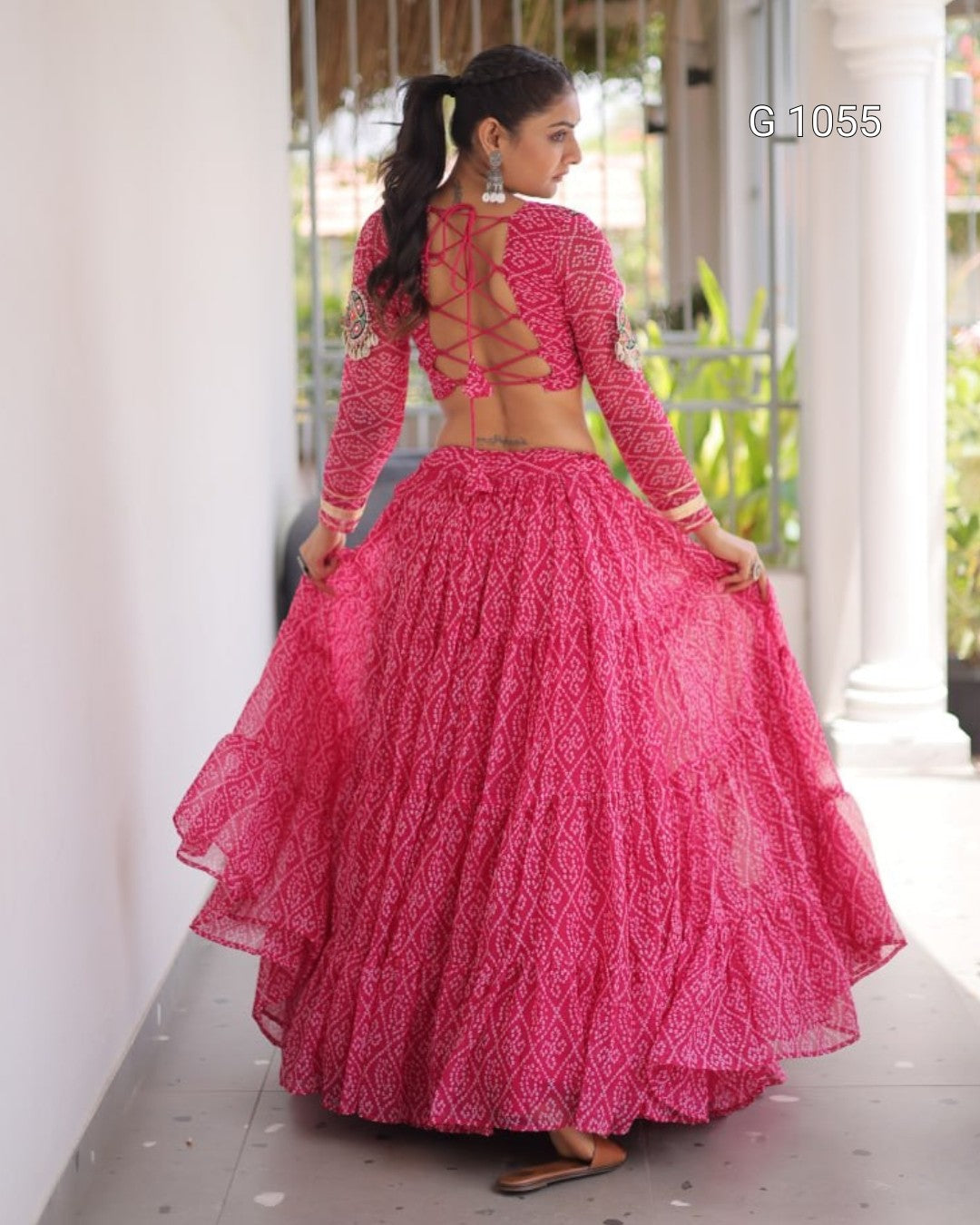 A woman wearing a bright pink embroidered traditional outfit with a long-sleeve lace choli and a full layered skirt, shown from the back in an outdoor corridor.