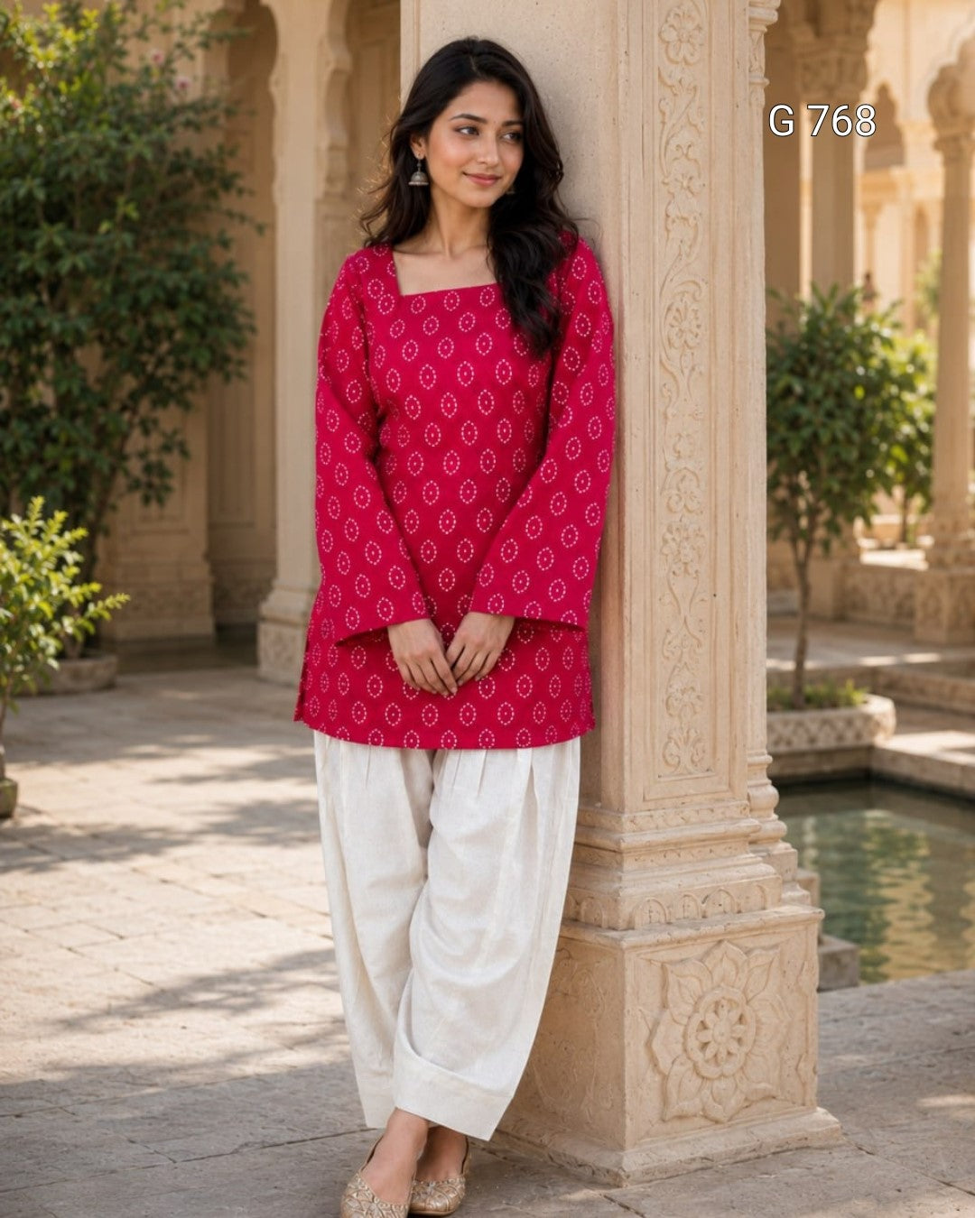 Woman in a red patterned top and white pants standing against an architectural column.