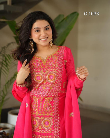 A woman wearing a bright pink traditional embroidered kurta set with matching dupatta, gold jewelry, and a warm smile, set against a softly blurred indoor background with green plants.