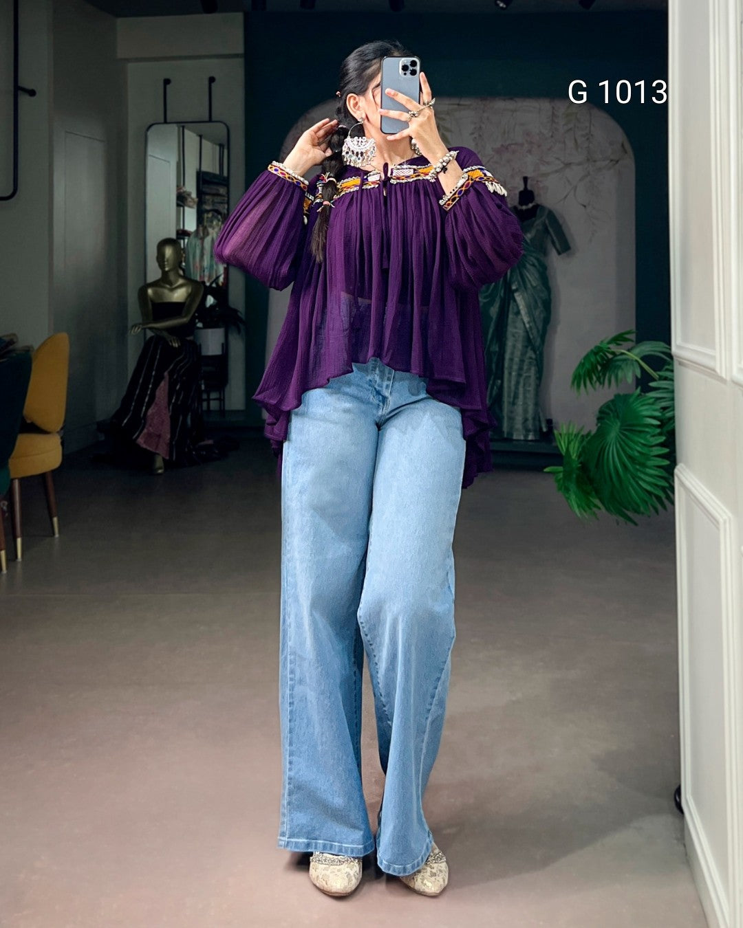 A woman takes a mirror selfie wearing a purple long-sleeve blouse with gold trim and light blue wide-leg jeans in a boutique setting.